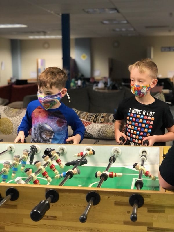 Two boys wearing face masks and playing foosball in a living room