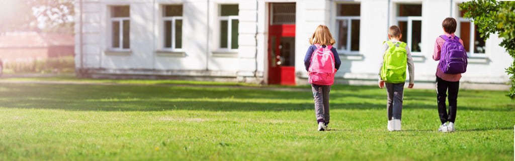 A group of kids walking towards a building in the grass with backpacks on
