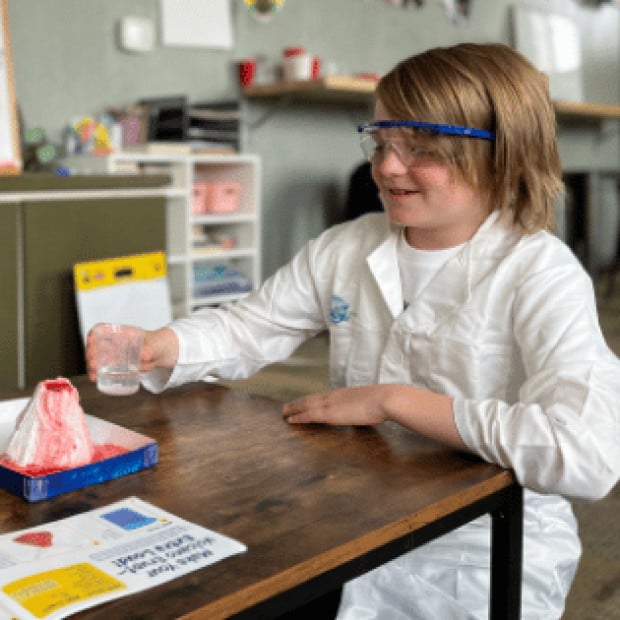 A young boy about to poor water into a volcano in a science classroom wearing a lab coat and glasses