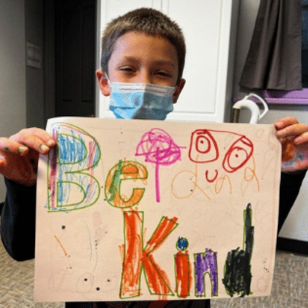 A young boy wearing a fast mask and holding up a hand drawn picture of the words 'Be Kind'