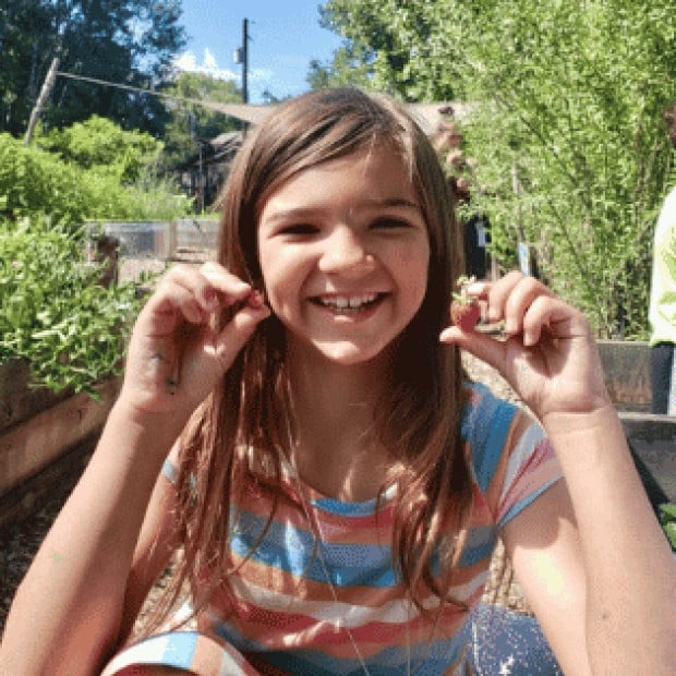 A young girl holding up fruit in front of a garden smiling 